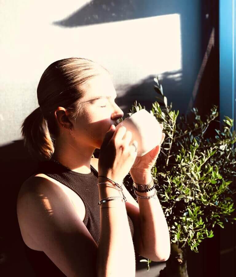 Eine Frau mit blondem, zusammengebundenen Haaren trinkt aus einer weißen Tasse. Sie trägt ein schwarzes Oberteil. Sonnenlicht scheint auf ihr Gesicht. Neben ihr steht eine grüne Pflanze.