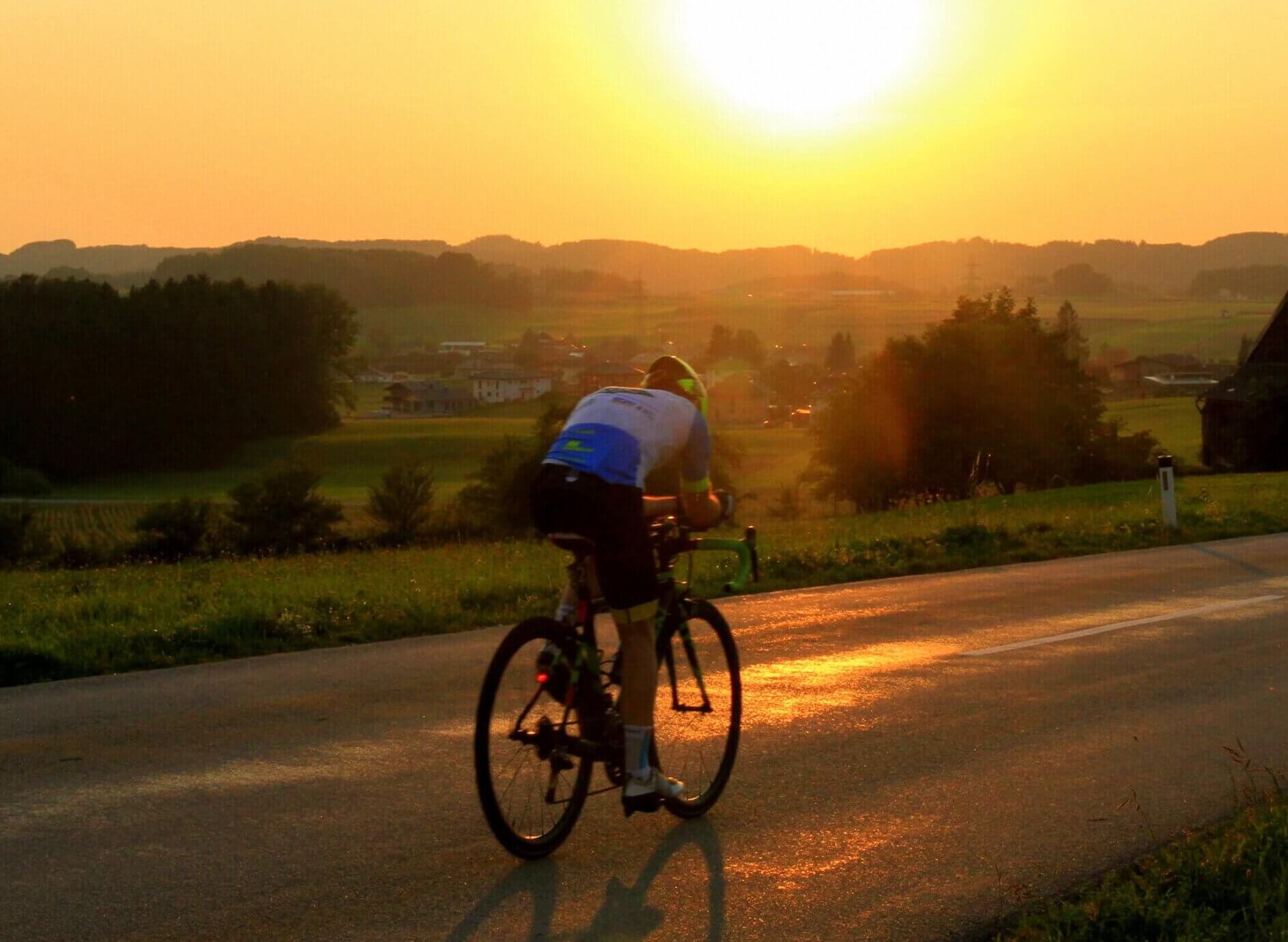 Das Bild zeigt einen Radsportler – Thomas Jaklitsch – der auf einem Rennrad eine Landstraße entlangfährt. Die Sonne steht tief am Horizont, taucht die Landschaft in goldenes Licht. Im Hintergrund sind sanfte Hügel, Bäume und ein ländliches Panorama zu sehen. Die Szene vermittelt Kraft, Ausdauer und den Moment der völligen Fokussierung. Das Bild steht im Kontext seiner Geschichte als Ultraradsportler, Mentalcoach und Speaker, der hajoona-Produkte gezielt für Energie, Erholung und Balance im Wettkampfalltag nutzt.